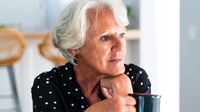 An elderly woman with short gray hair and a black polka dot shirt sits holding a mug, gazing thoughtfully to the side in a bright, cozy room.