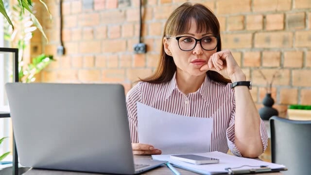 A woman wearing glasses sits at a desk with a laptop, holding papers and looking thoughtful. There are notebooks, a phone, and a pen on the desk. She is in a bright room with a brick wall background.