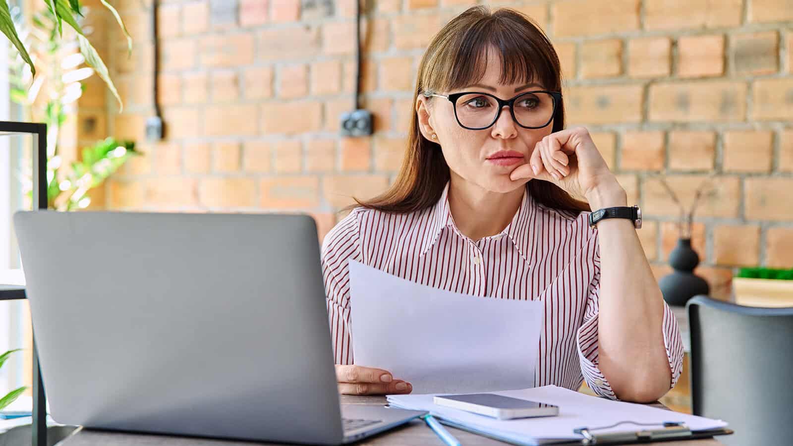 A woman wearing glasses sits at a desk with a laptop, holding papers and looking thoughtful. There are notebooks, a phone, and a pen on the desk. She is in a bright room with a brick wall background.