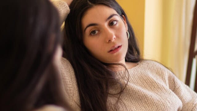 A woman with long dark hair, wearing a beige sweater, looks thoughtfully at her reflection in a mirror. Warm yellow and cream tones fill the background.