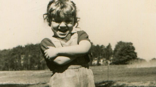 A young child smiles brightly with arms crossed, standing outdoors in a field. Trees and a clear sky are visible in the blurred background. The photo is black and white, giving it a vintage feel.