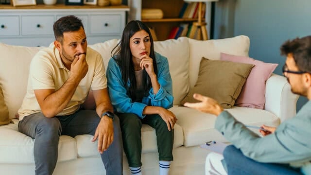 A couple sits on a couch, looking concerned and listening to a person across from them who appears to be a counselor or therapist, gesturing while speaking.