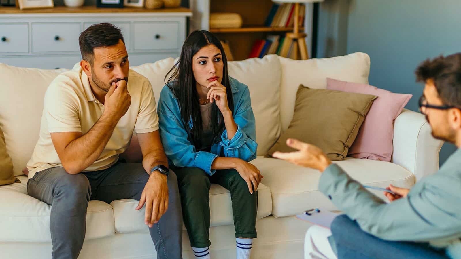 A couple sits on a couch, looking concerned and listening to a person across from them who appears to be a counselor or therapist, gesturing while speaking.
