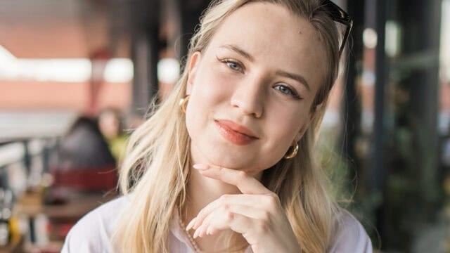 A woman with long blonde hair and gold hoop earrings smiles softly, resting her chin on her hand, sitting outdoors at a cafe with blurred people and tables in the background.
