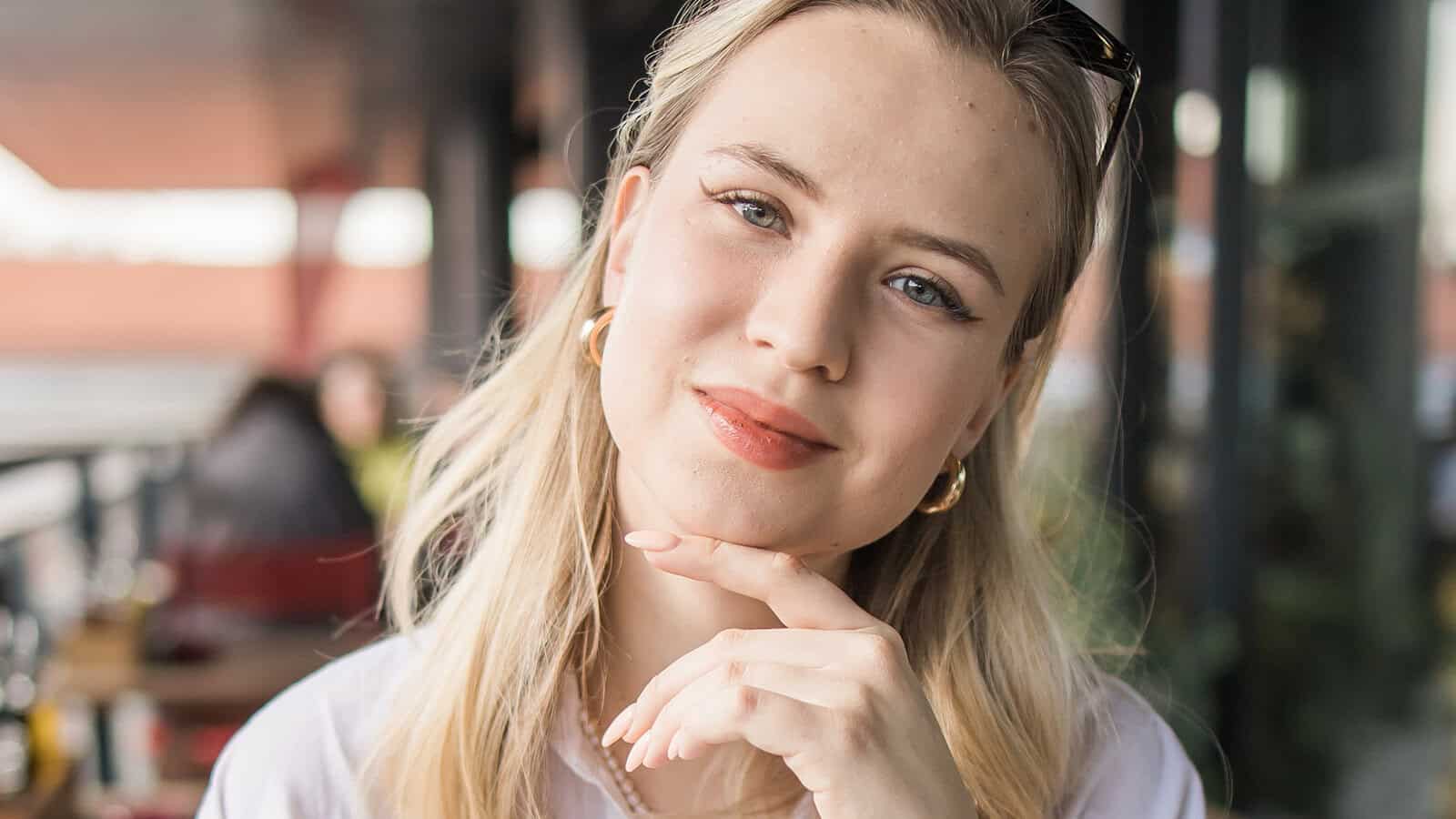 A woman with long blonde hair and gold hoop earrings smiles softly, resting her chin on her hand, sitting outdoors at a cafe with blurred people and tables in the background.