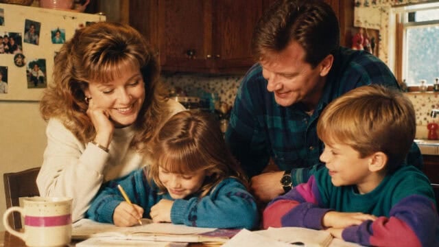 A smiling millennial family of four sits at a kitchen table, parents watching as a young girl writes in a notebook, while a boy beside her laughs. Warm, homey kitchen setting with a mug and papers on the table adds to the cozy scene, echoing memories of boomer parents sharing old-fashioned phrases over breakfast.