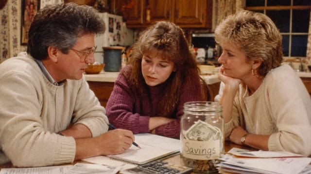 A man, young woman, and older woman sit at a kitchen table discussing papers. A jar labeled Savings with money inside and a calculator are on the table. The kitchen background is warmly lit and cozy.