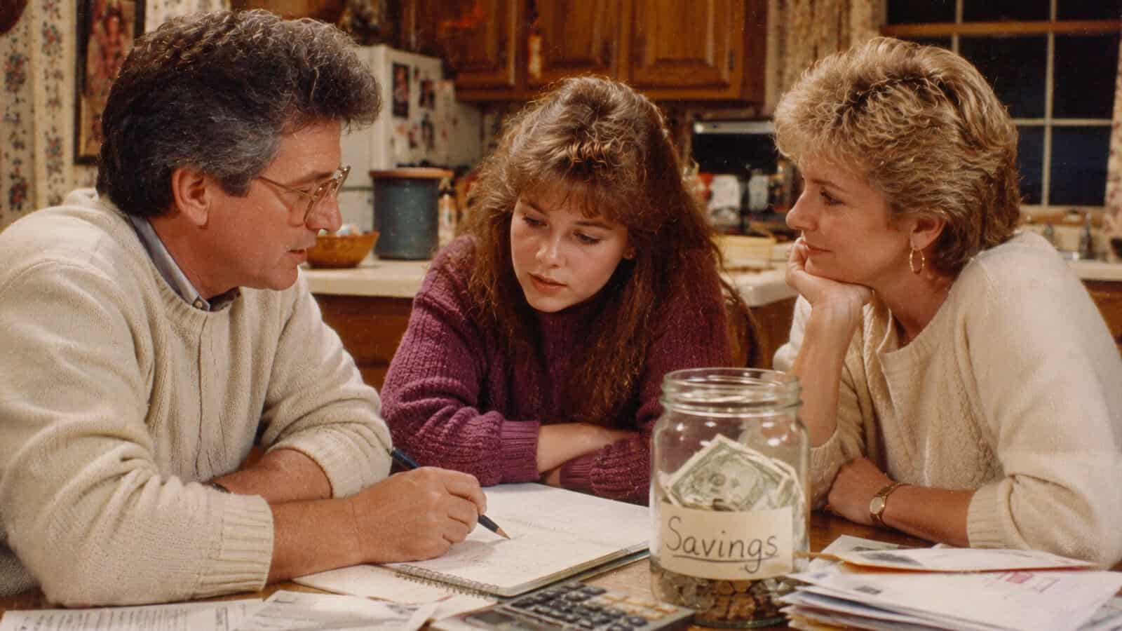 A man, young woman, and older woman sit at a kitchen table discussing papers. A jar labeled Savings with money inside and a calculator are on the table. The kitchen background is warmly lit and cozy.