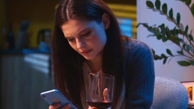 A woman with long dark hair looks at her smartphone while holding a glass of red wine, sitting indoors next to a plant.
