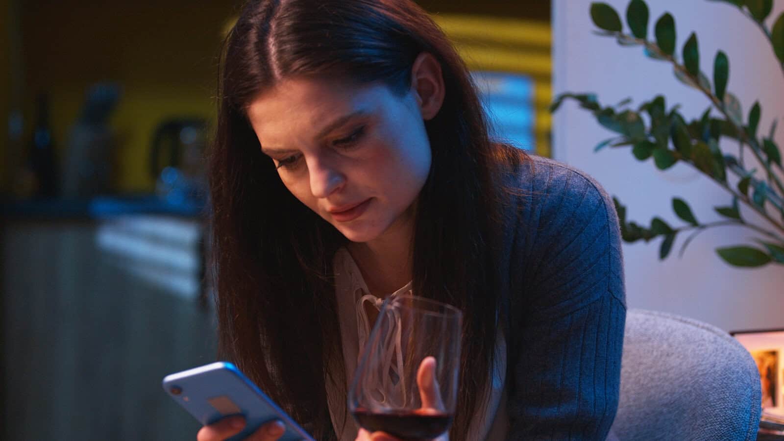 A woman with long dark hair looks at her smartphone while holding a glass of red wine, sitting indoors next to a plant.