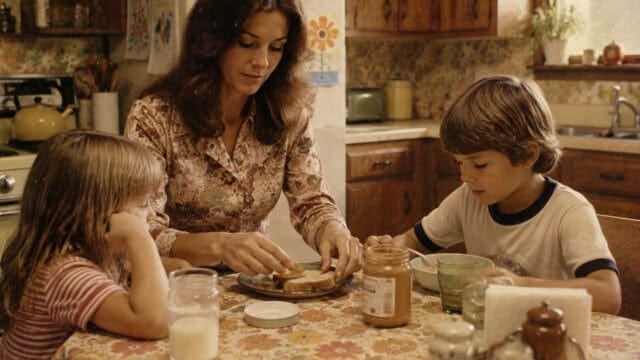 A woman prepares peanut butter sandwiches for two children at a kitchen table. The table is set with milk, a jar of peanut butter, and plates. The kitchen has warm, vintage decor with wood cabinets and floral wallpaper.