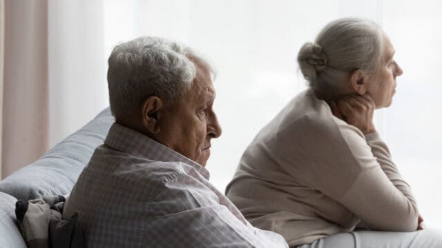 Elderly man and woman sit on a couch, facing away from each other, both looking pensive or upset by a window with soft daylight.