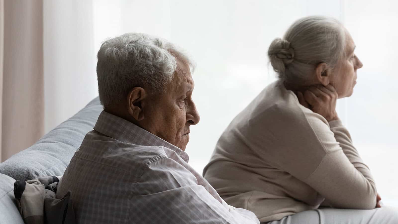 Elderly man and woman sit on a couch, facing away from each other, both looking pensive or upset by a window with soft daylight.