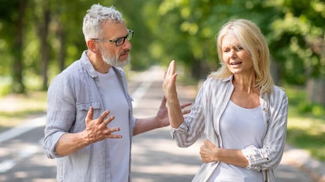 An older man and woman argue on a tree-lined path. The man gestures with open hands, looking frustrated. The woman raises her hand as if to stop him and looks upset, turning away from him as they stand close together outdoors.