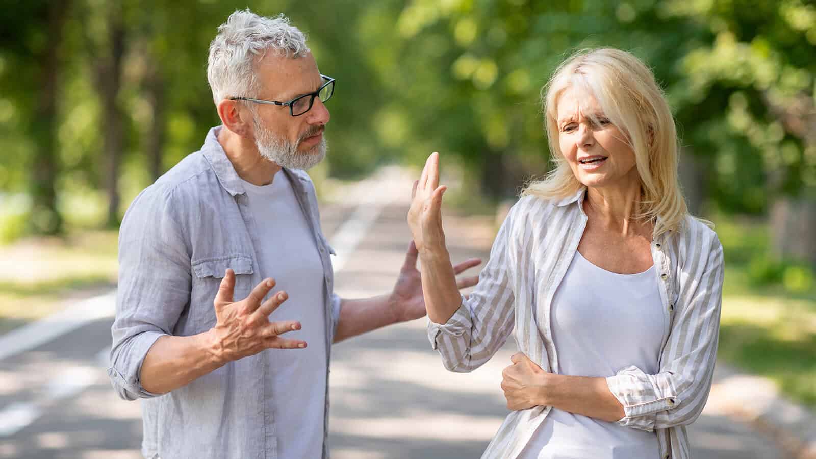 An older man and woman argue on a tree-lined path. The man gestures with open hands, looking frustrated. The woman raises her hand as if to stop him and looks upset, turning away from him as they stand close together outdoors.