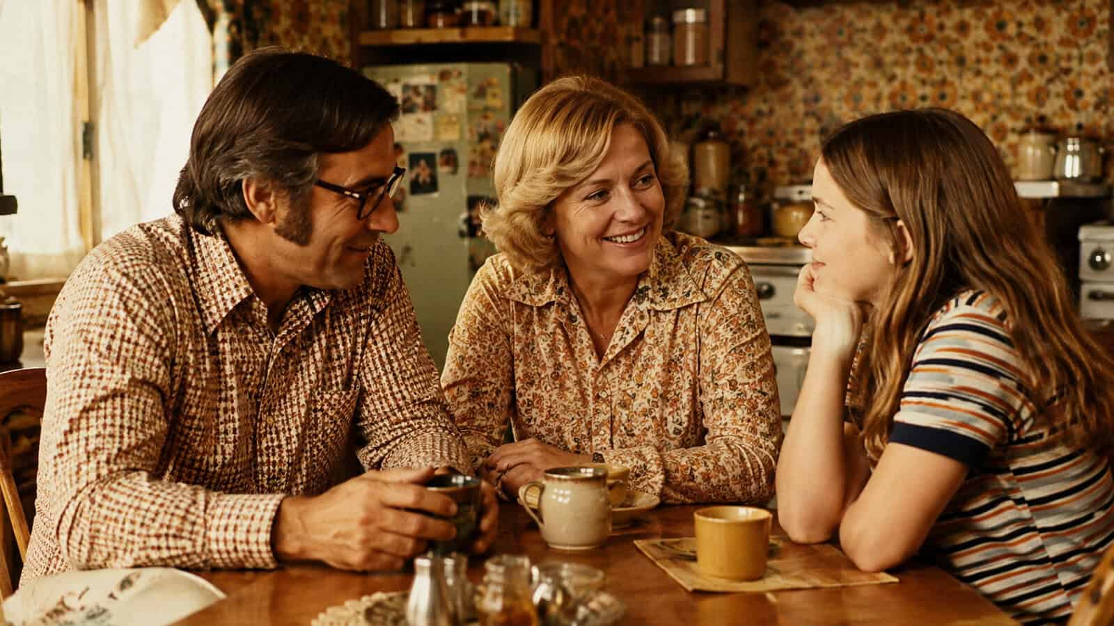 Three people sit at a kitchen table, smiling and talking. The adults wear patterned shirts, the girl wears stripes. The kitchen has a vintage look with floral wallpaper, an old fridge, and various mugs on the table.
