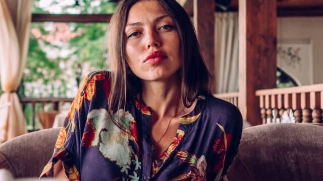 A woman with long brown hair wearing a colorful floral shirt sits indoors on a cushioned chair, looking directly at the camera with a neutral expression. Wooden beams and greenery are visible in the background.