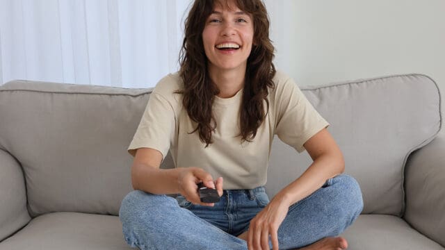 A smiling person with wavy brown hair sits cross-legged on a light gray couch, holding a remote control and wearing a beige t-shirt and blue jeans. White curtains are visible in the background.