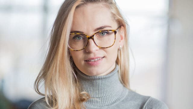 A woman with long blonde hair and tortoiseshell glasses wears a gray turtleneck sweater, smiling softly with a blurred background.