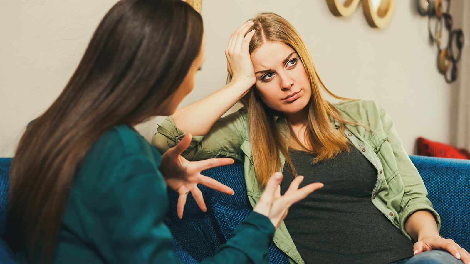 Two women sit on a couch. One gestures while talking, and the other looks frustrated, resting her head on her hand and listening.