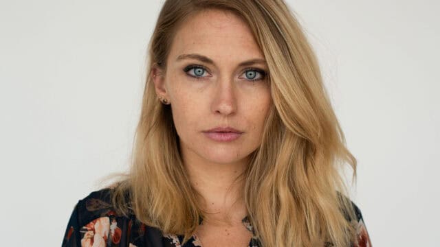 A woman with long blonde hair and blue eyes stares directly at the camera. She is wearing a dark floral top and small earrings, with a neutral expression and a plain light background.