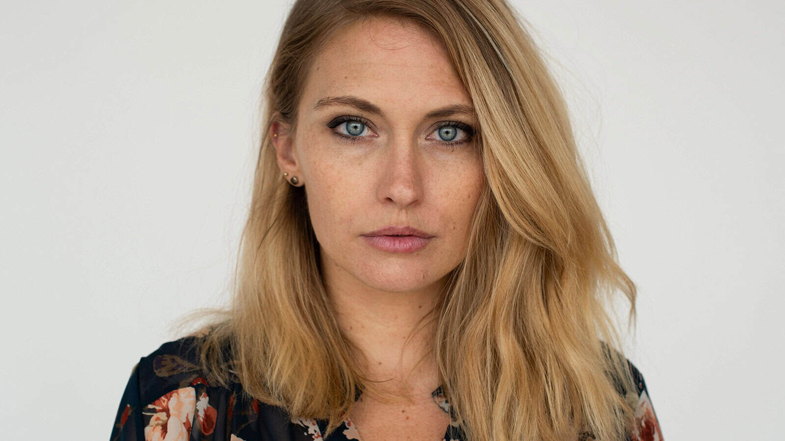 A woman with long blonde hair and blue eyes stares directly at the camera. She is wearing a dark floral top and small earrings, with a neutral expression and a plain light background.