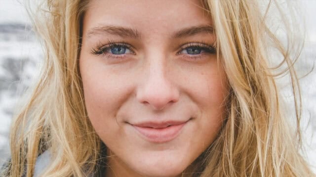A close-up of a young woman with long blonde hair, blue eyes, and a slight smile, standing outdoors with a blurred background.