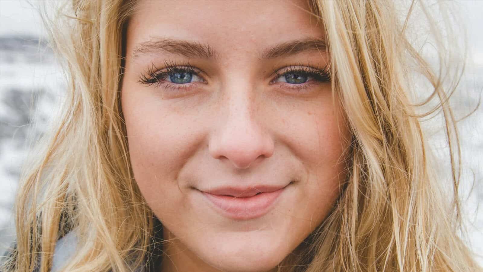 A close-up of a young woman with long blonde hair, blue eyes, and a slight smile, standing outdoors with a blurred background.