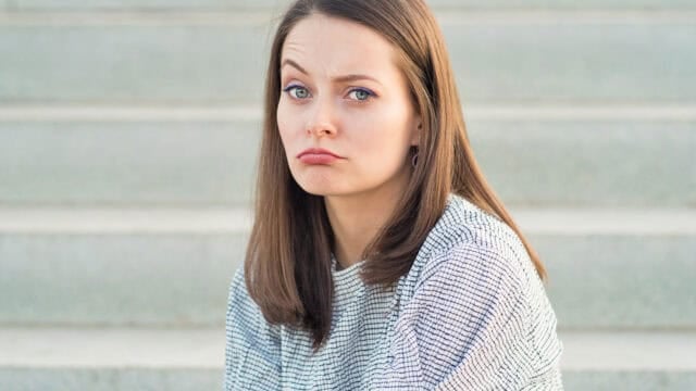A woman with straight brown hair is sitting on outdoor steps, wearing a white and black patterned sweater, and making a sad pouty face while looking at the camera.