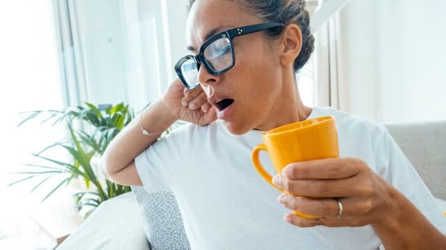 Woman wearing glasses yawns while holding a yellow mug, sitting on a couch in a bright room with plants in the background.