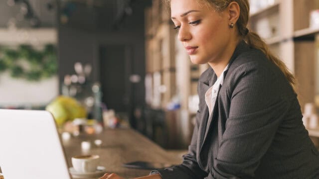 A woman in a gray blazer works on a laptop at a wooden counter in a cafe, with a cup of coffee nearby and shelves blurred in the background.