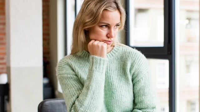 A woman in a mint green sweater sits indoors, resting her chin on her fist and looking pensively to the side by a window.