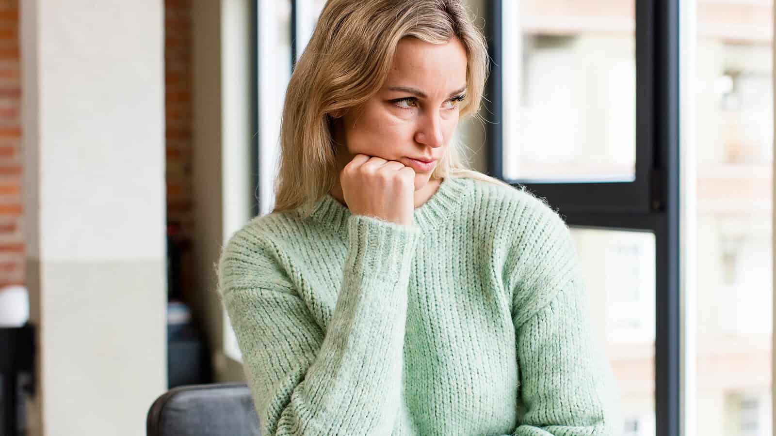 A woman in a mint green sweater sits indoors, resting her chin on her fist and looking pensively to the side by a window.