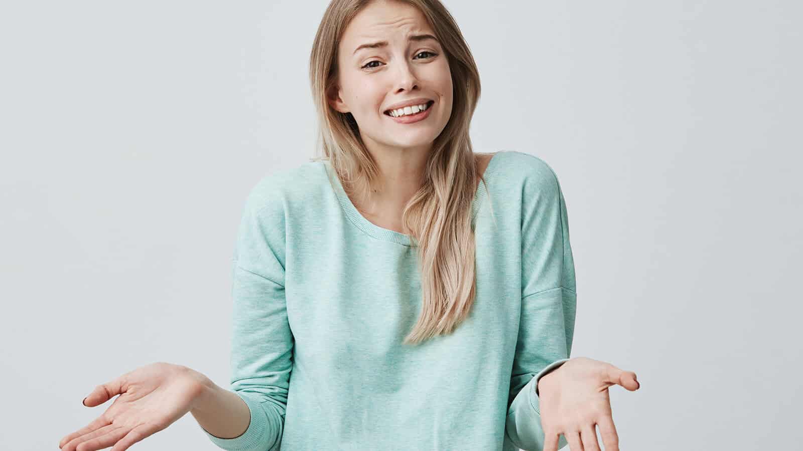 A woman with long blonde hair wearing a light blue sweater shrugs her shoulders with raised eyebrows and upturned palms, expressing confusion or uncertainty, against a plain light background.