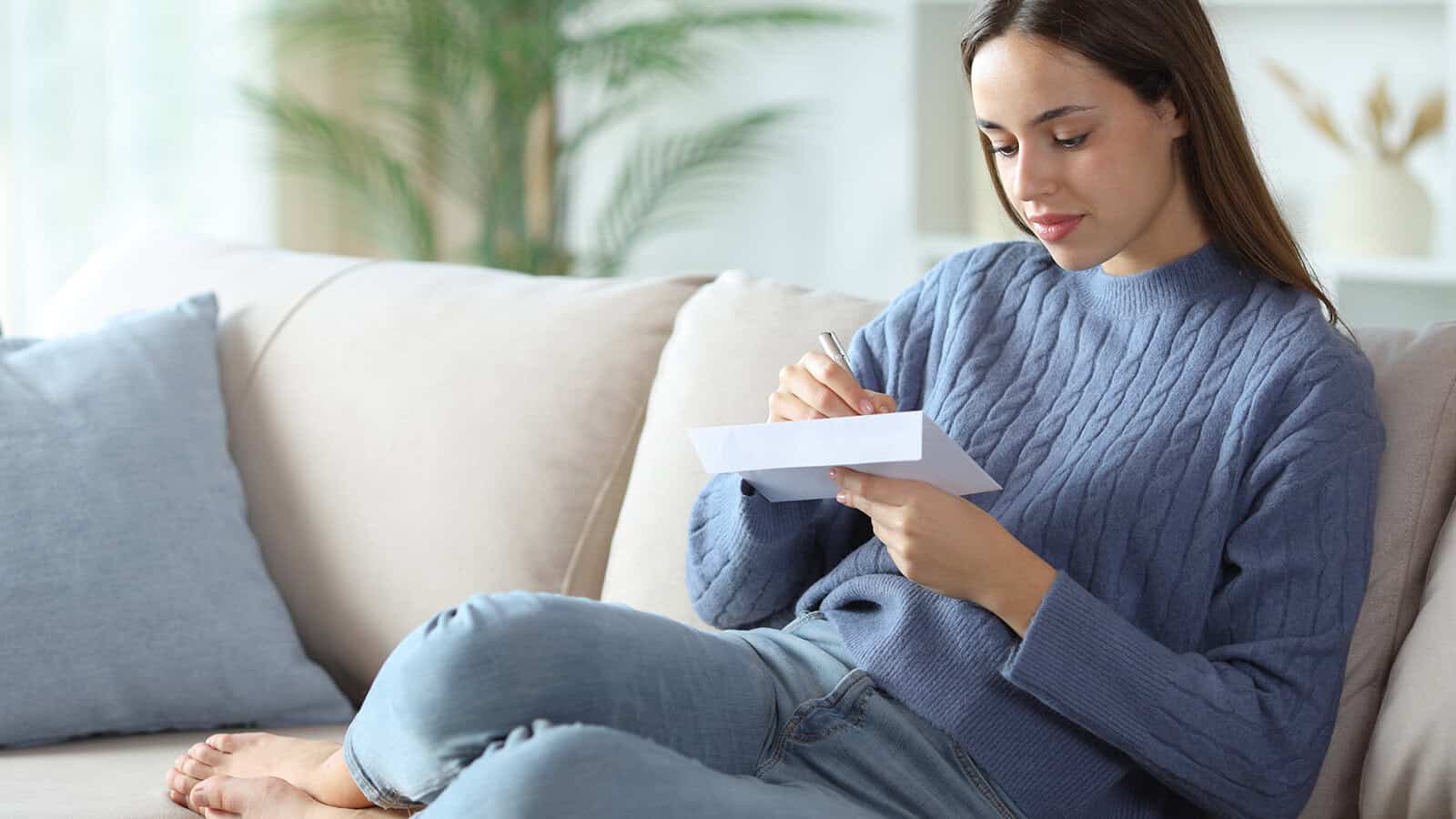 A woman in a blue sweater and jeans sits on a couch, writing on a piece of paper, with a pillow and plants in the background.