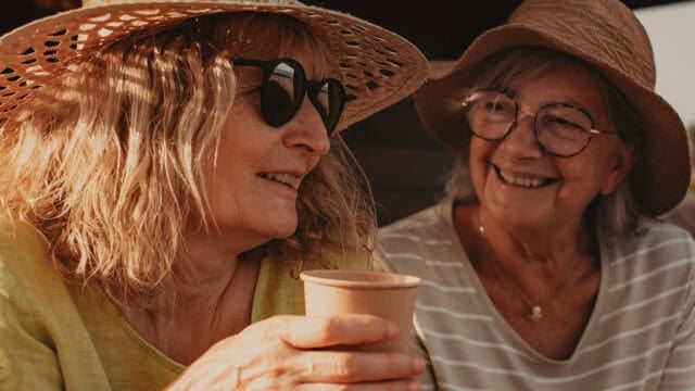 Two older women wearing sun hats and glasses smile at each other outdoors. One holds a paper cup, and both appear relaxed and happy, bathed in warm sunlight.