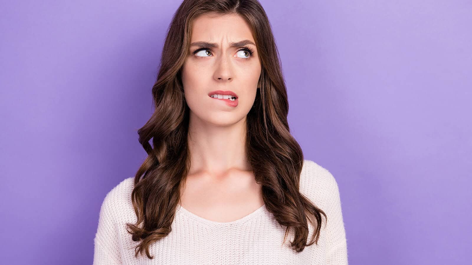 A woman with long brown hair, wearing a white sweater, stands against a purple background, looking to the side with a worried or uncertain expression while biting her lip.