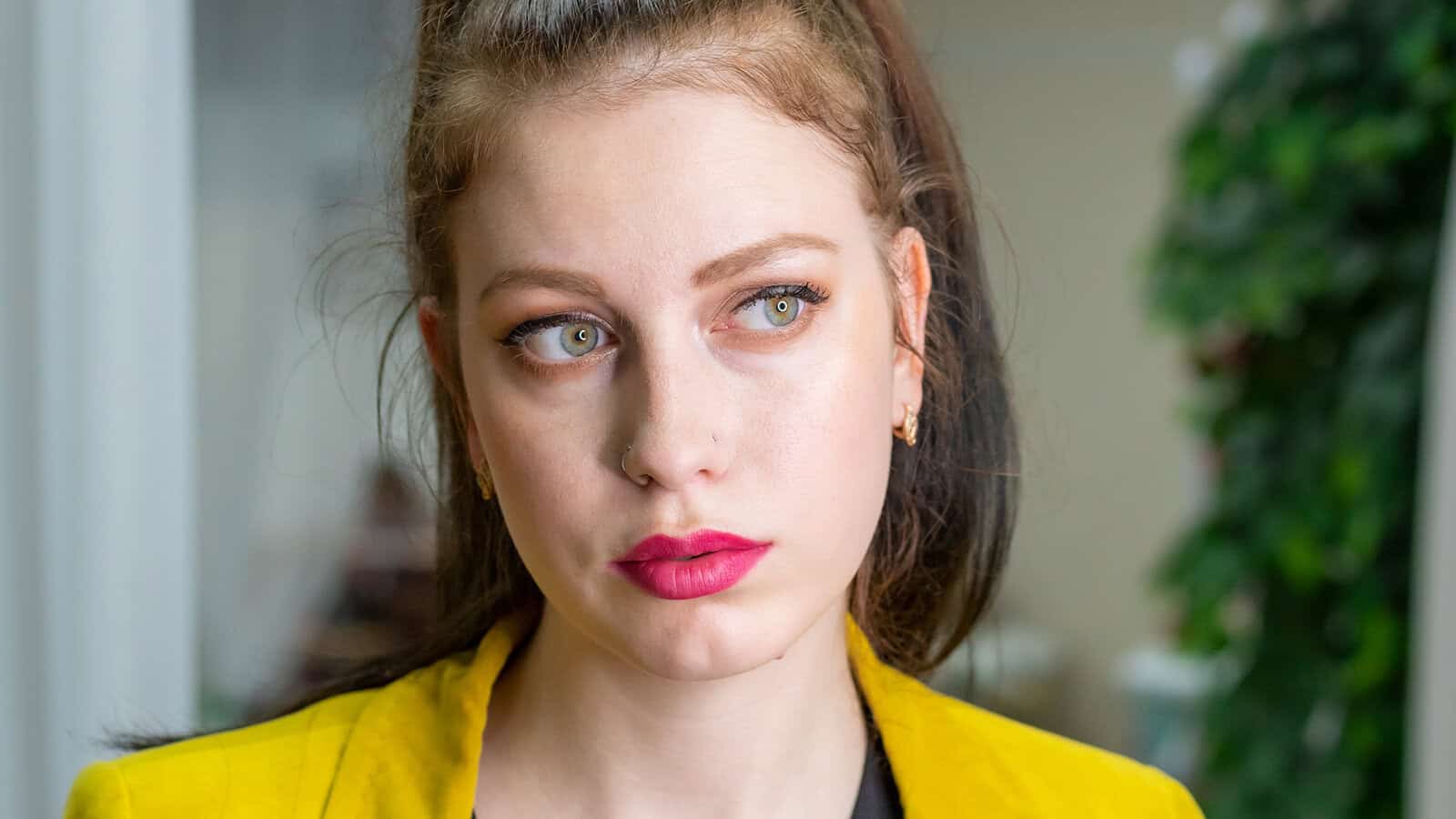 Young woman with long brown hair and bright pink lipstick, wearing a yellow jacket, looks to the side with a serious expression. The background is blurred with greenery and light-colored curtains visible.
