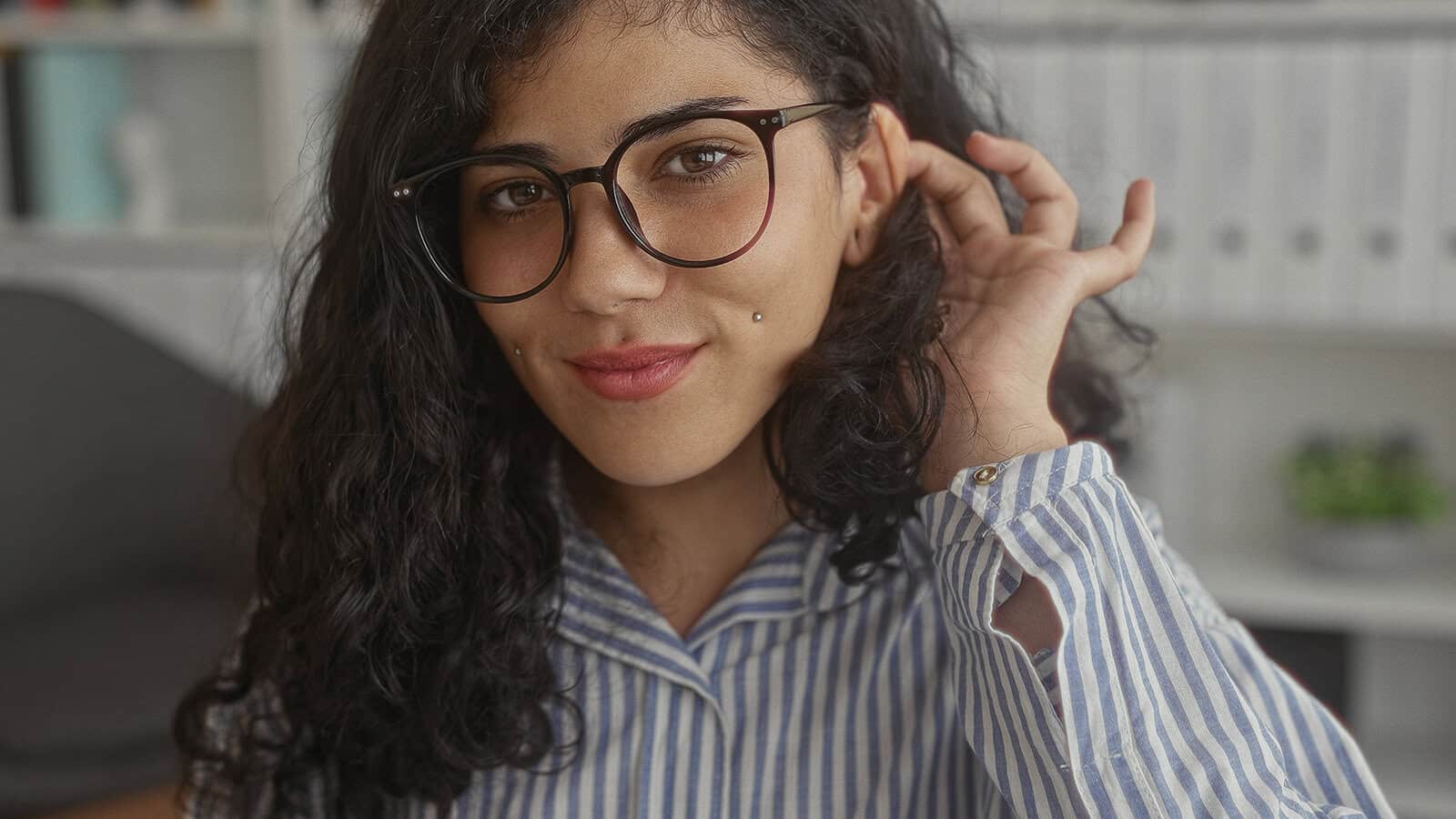A person with long curly hair, glasses, and a striped shirt smiles softly at the camera, touching their hair. The background is softly blurred, showing shelves and binders.
