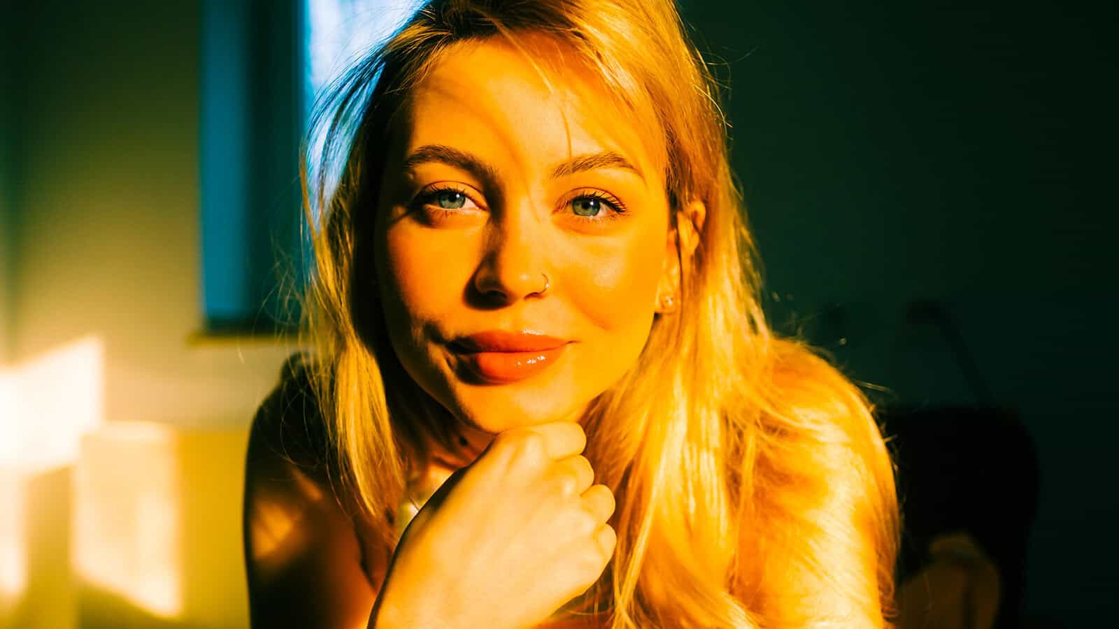 A woman with blonde hair sits indoors, bathed in warm golden sunlight. She rests her chin on her hand and looks into the camera, with a window and dark background behind her.