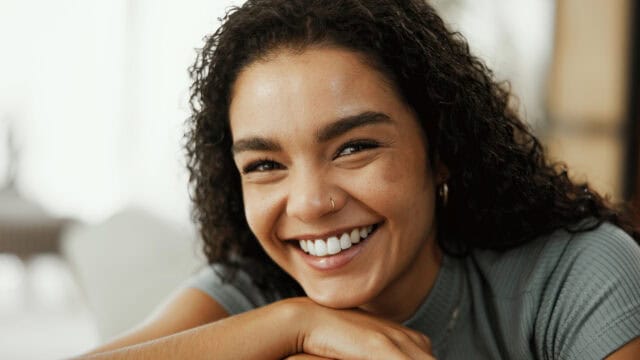 A young woman with curly hair and a nose ring smiles brightly, resting her chin on her hands indoors with soft lighting.