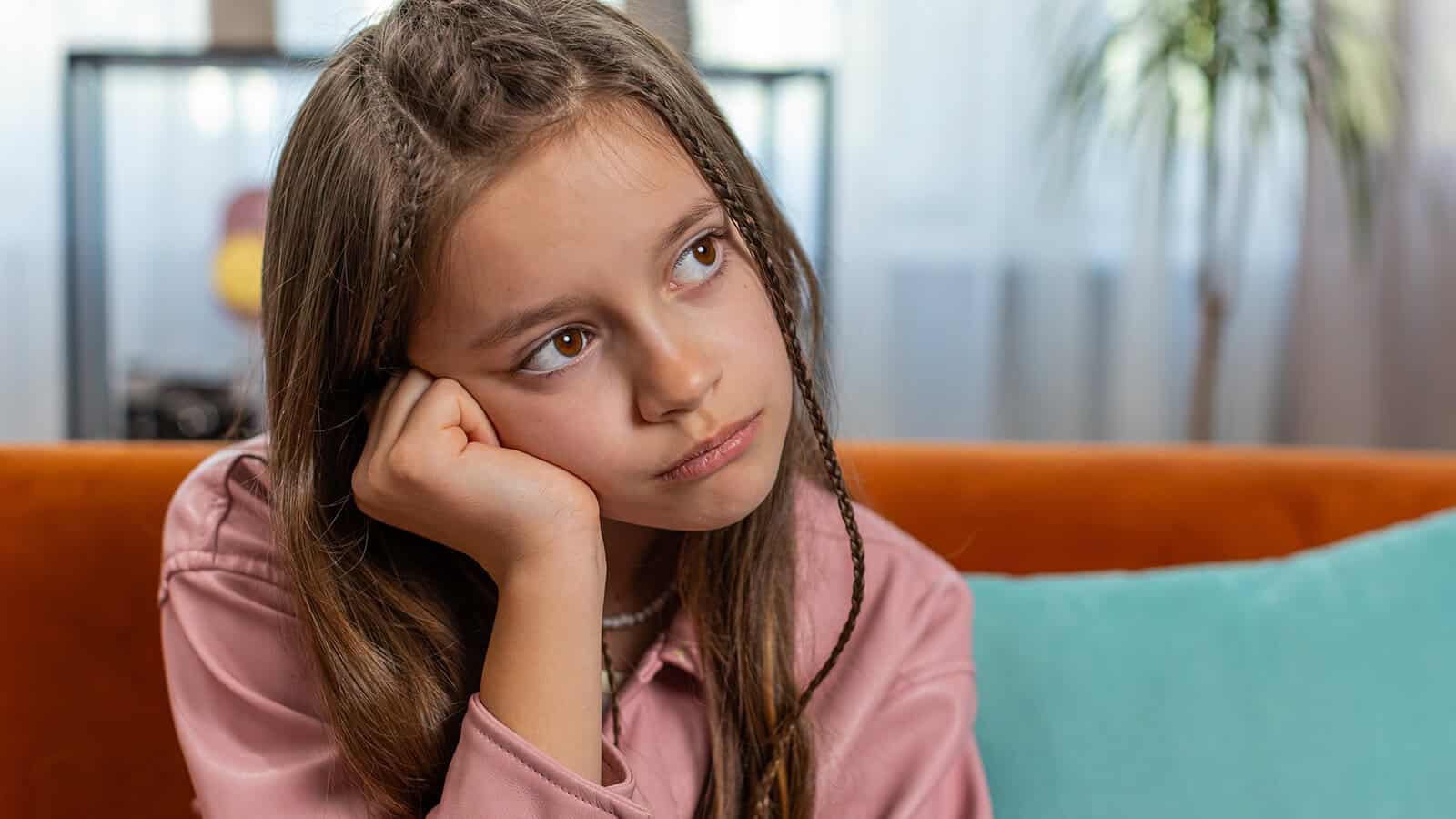 A young girl with long brown hair sits on an orange couch, resting her head on her hand and looking off to the side with a thoughtful, bored expression. A turquoise pillow and blurred background are visible.