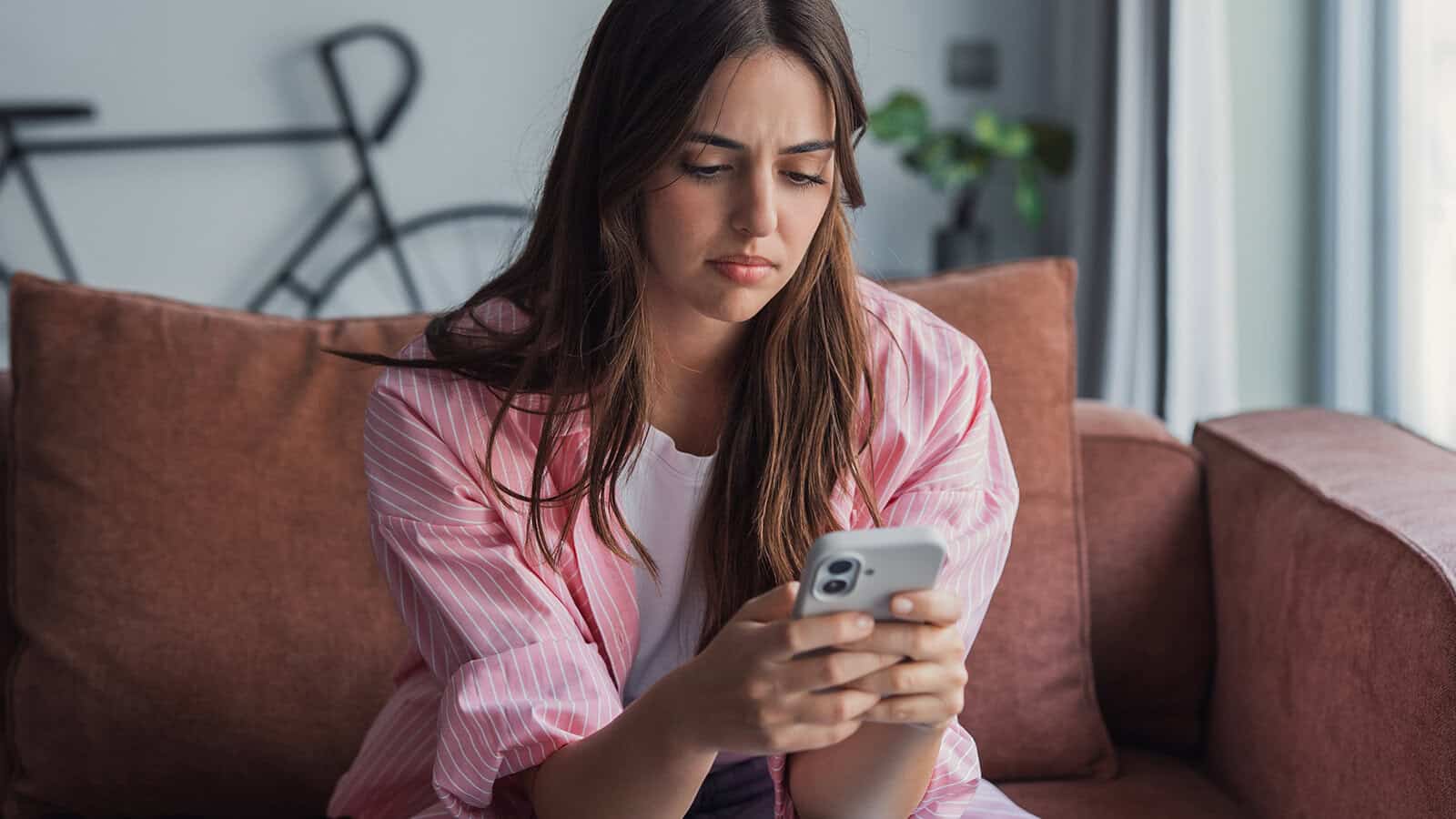 A young woman sits on a couch looking concerned while reading something on her smartphone, with a bicycle and plant in the background.