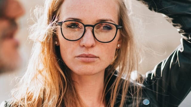 A woman with long blonde hair and black glasses looks ahead with a serious expression, wearing a dark jacket. Sunlight highlights her face and hair.