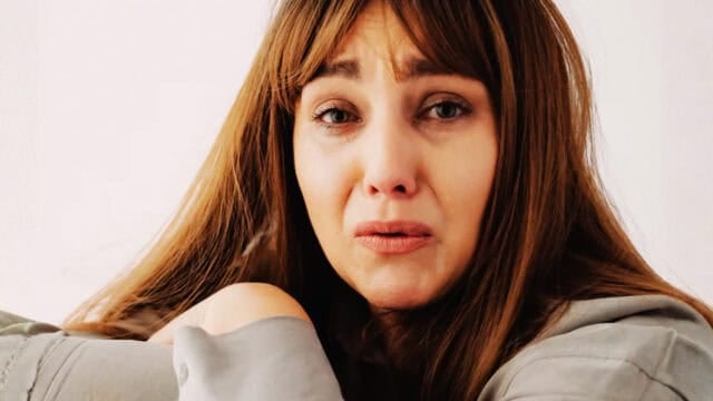 A woman with long brown hair looks distressed and teary-eyed, holding her knee close to her chest while wearing a gray top, against a plain light background.