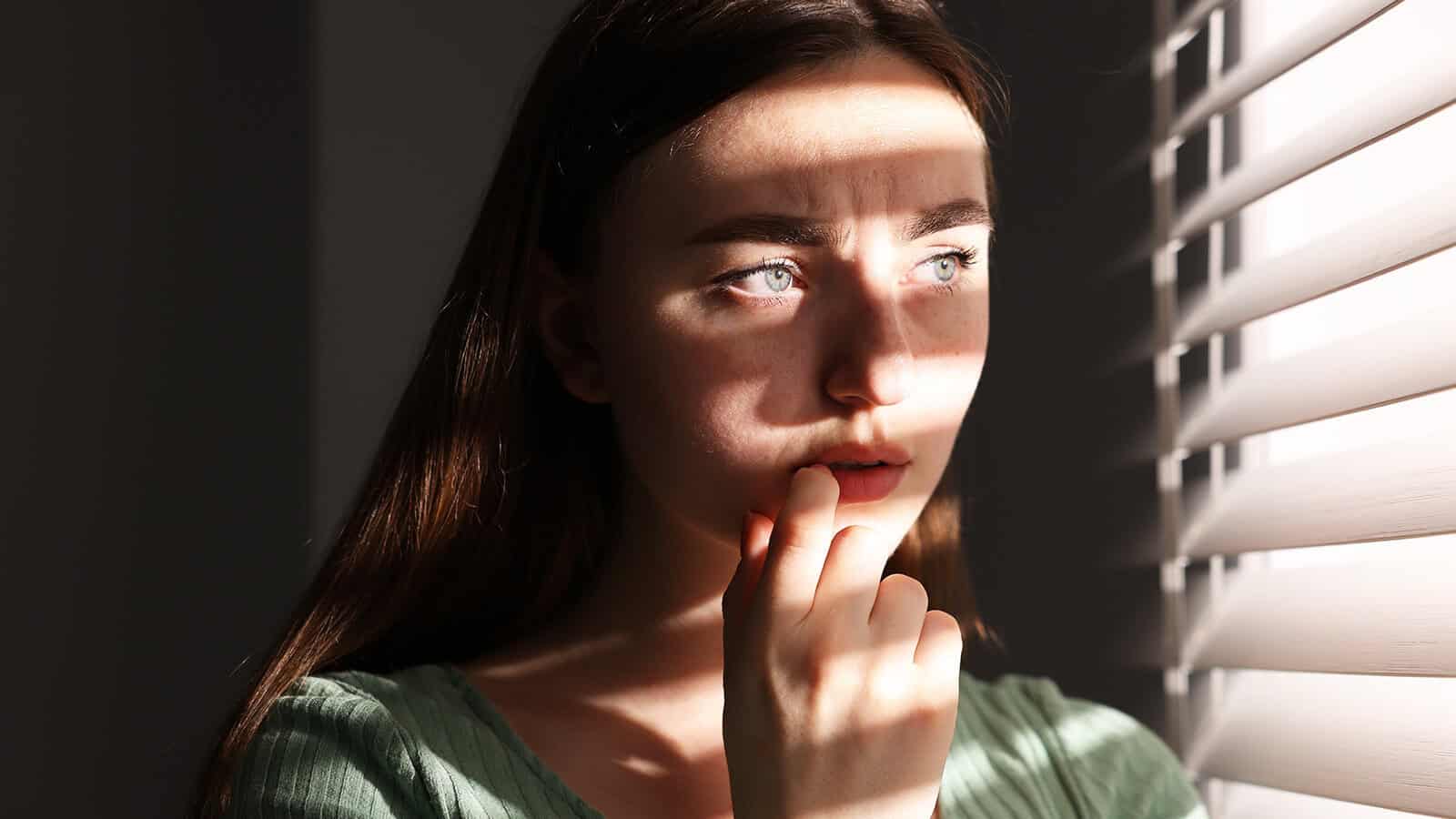 A woman with long brown hair stands near window blinds, sunlight casting striped shadows on her face. She looks thoughtful, with her hand touching her lips.