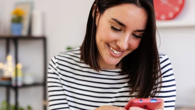 A woman wearing a black-and-white striped shirt smiles while looking at her smartphone. There is a shelf with plants and a red clock in the blurred background.