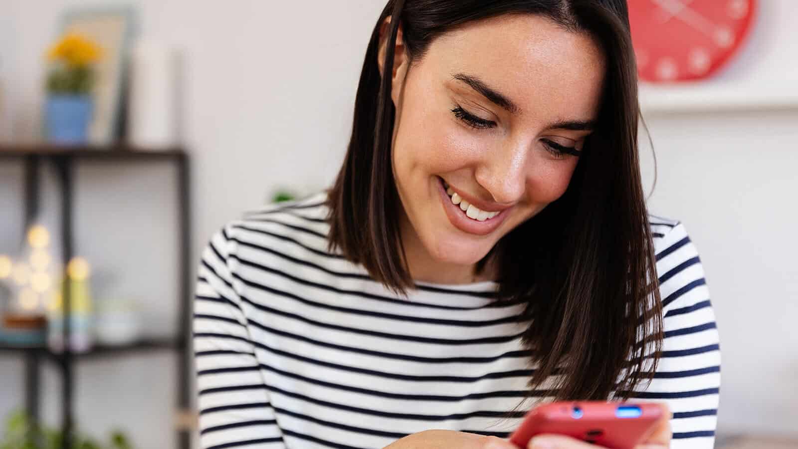 A woman wearing a black-and-white striped shirt smiles while looking at her smartphone. There is a shelf with plants and a red clock in the blurred background.