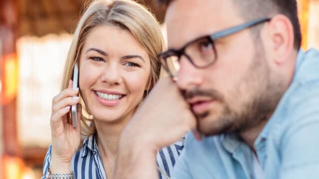 A smiling woman talks on a phone while a man with glasses sits nearby, appearing thoughtful or distracted. They are outdoors in a casual setting.
