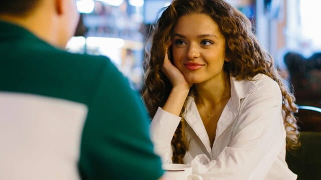 A woman with curly hair, wearing a white shirt, sits at a table in a café, resting her head on her hand and smiling while looking at someone across from her.